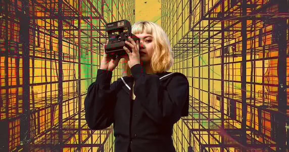 A woman with short blonde hair wearing a black dress takes a photo with a black camera on a yellow, green, and orange background with a geometric pattern on it.